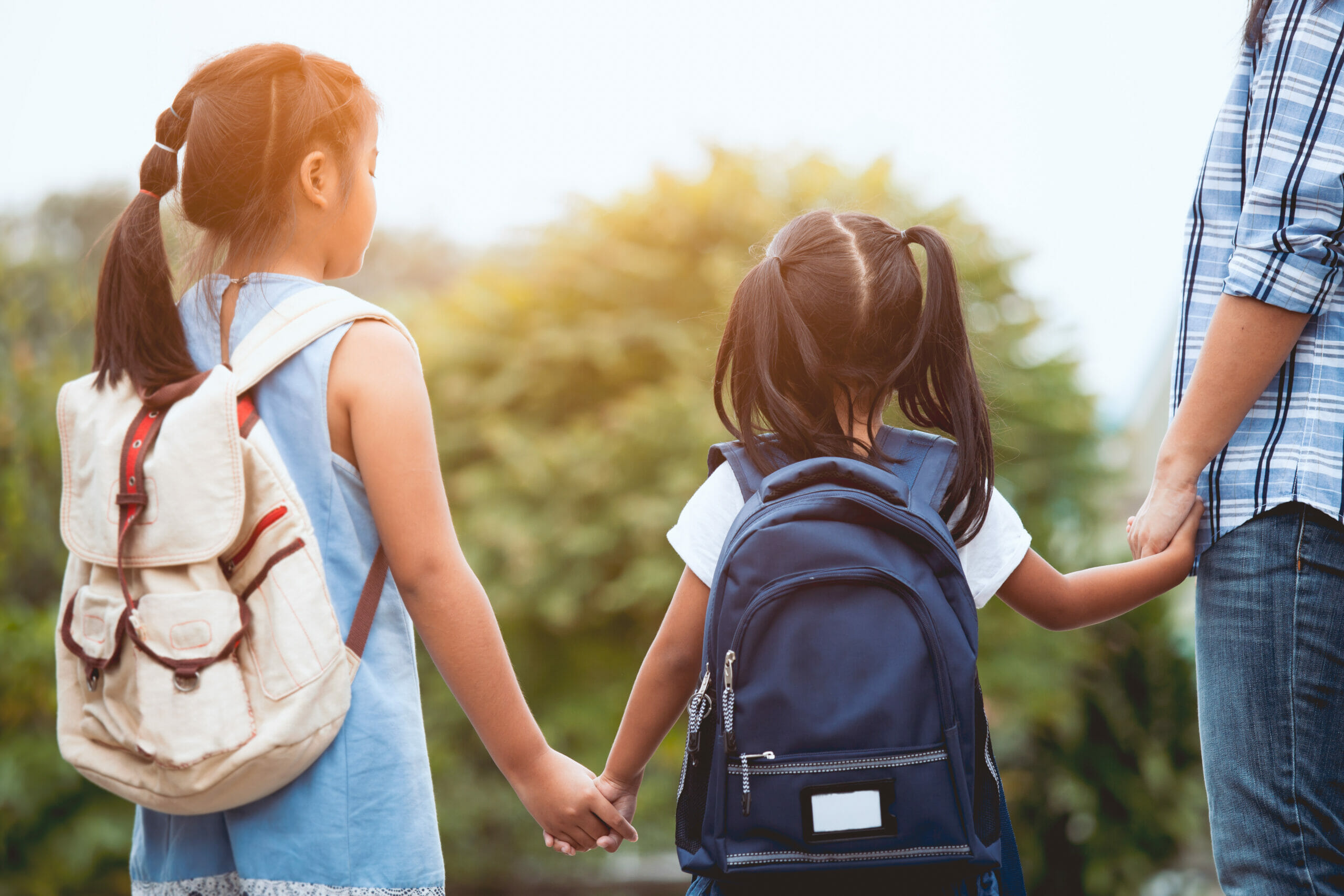 Two little girls wearing backpacks hold hands with an adult.